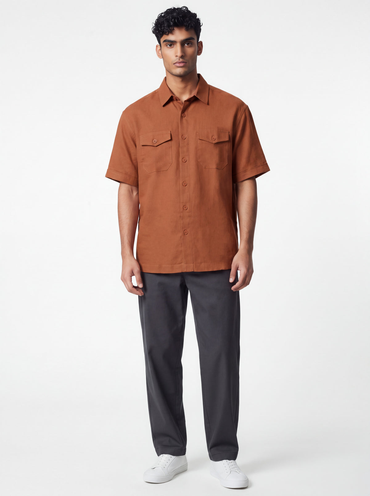 A man modeling a short-sleeve button-up shirt in rust orange, paired with dark gray trousers and white sneakers, against a plain light gray studio background.