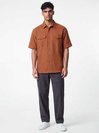 A man modeling a short-sleeve button-up shirt in rust orange, paired with dark gray trousers and white sneakers, against a plain light gray studio background.