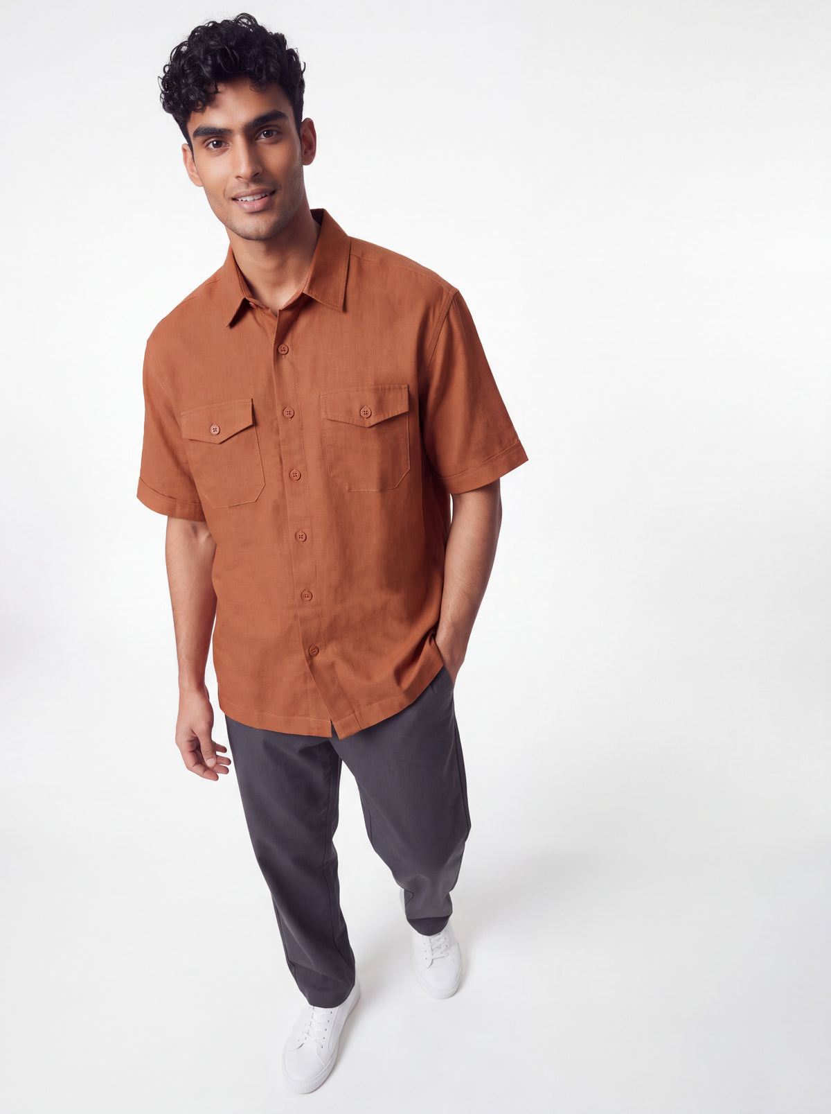 A man wearing a rust-orange short-sleeve button-up shirt with two chest pockets, paired with dark gray pants and white sneakers, standing against a plain white studio background.