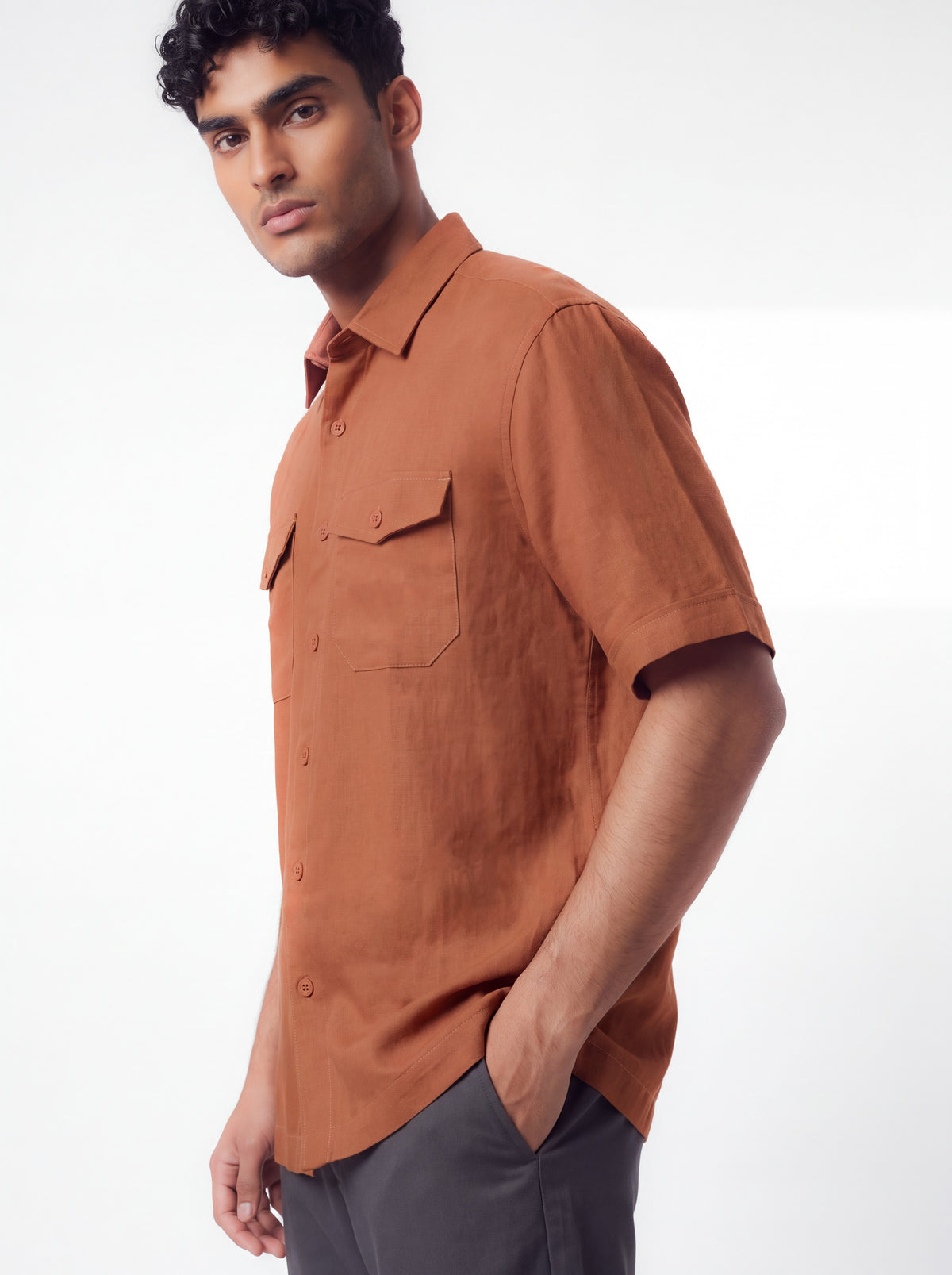 A man wearing a short-sleeve button-up shirt in a warm rust or terracotta color, paired with dark gray trousers, photographed against a clean white studio background.