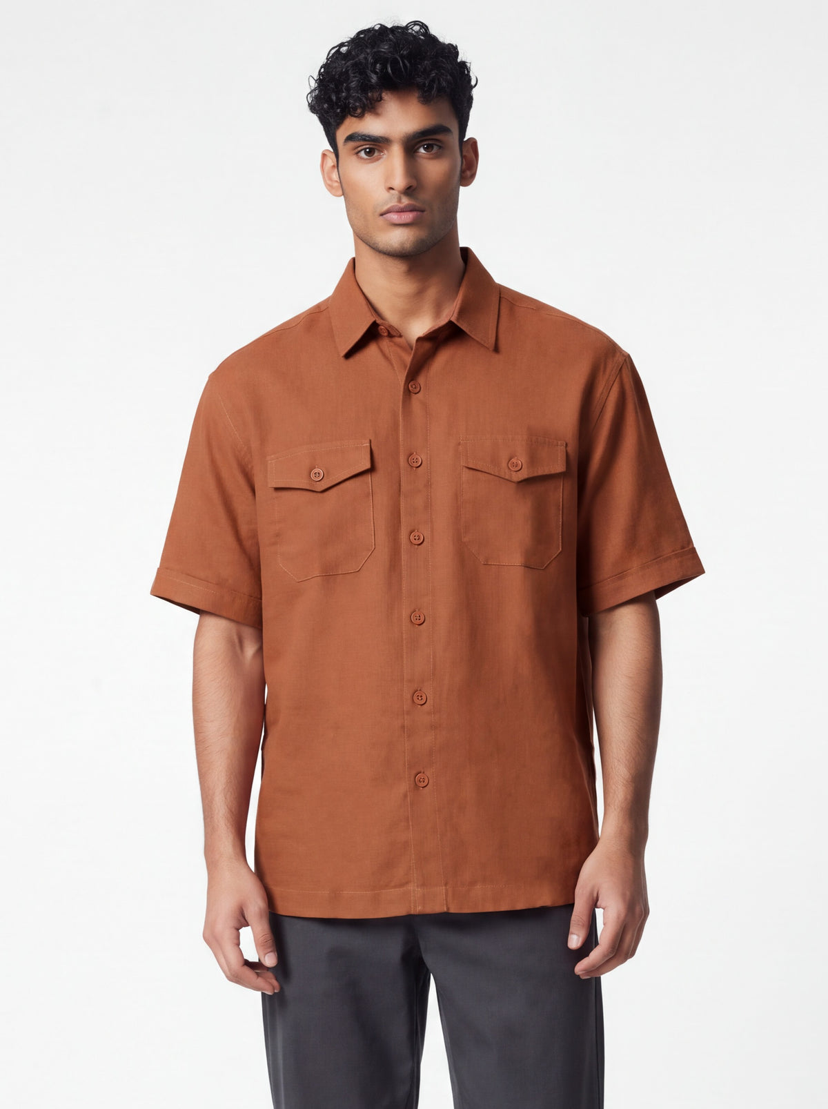 A man wearing a short-sleeve button-up shirt in a warm rust brown color, paired with dark gray trousers, standing against a plain white studio background.
