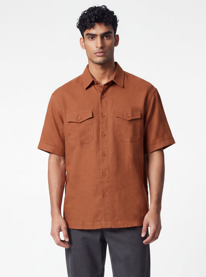 A man wearing a short-sleeve button-up shirt in a warm rust brown color, paired with dark gray trousers, standing against a plain white studio background.
