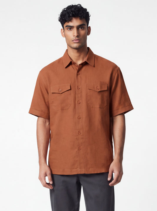 A man wearing a short-sleeve button-up shirt in a warm rust brown color, paired with dark gray trousers, standing against a plain white studio background.
