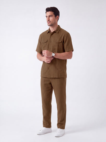 A man modeling a matching brown short-sleeve button-up shirt and trousers set, paired with white sneakers, against a plain light gray studio background.