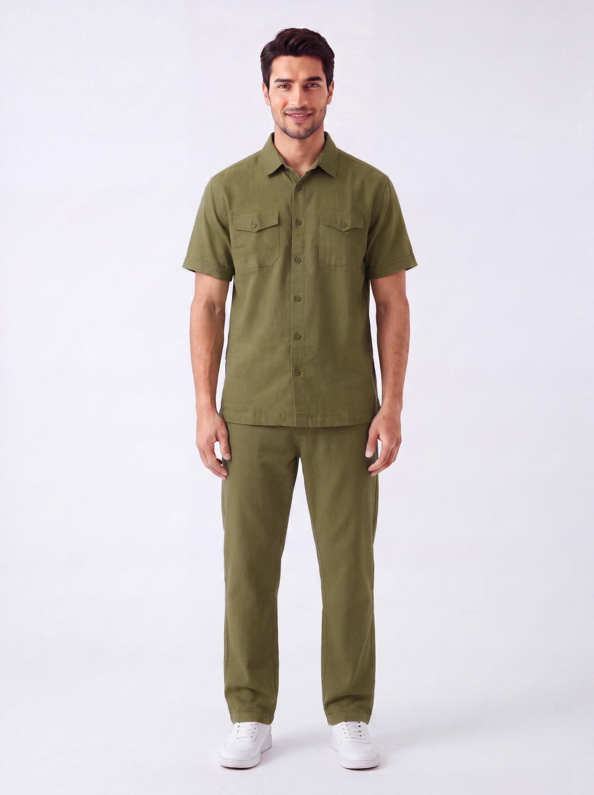 A man modeling a matching olive green short-sleeve button-up shirt and trousers set, paired with white sneakers, against a plain light gray studio background.