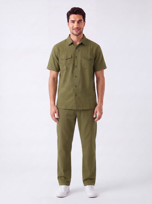A man modeling a matching olive green short-sleeve button-up shirt and trousers set, paired with white sneakers, against a plain light gray studio background.