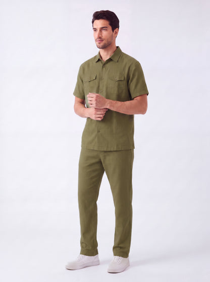 A man modeling a matching olive green short-sleeve button-up shirt and trousers set, paired with white sneakers, against a plain white studio background.