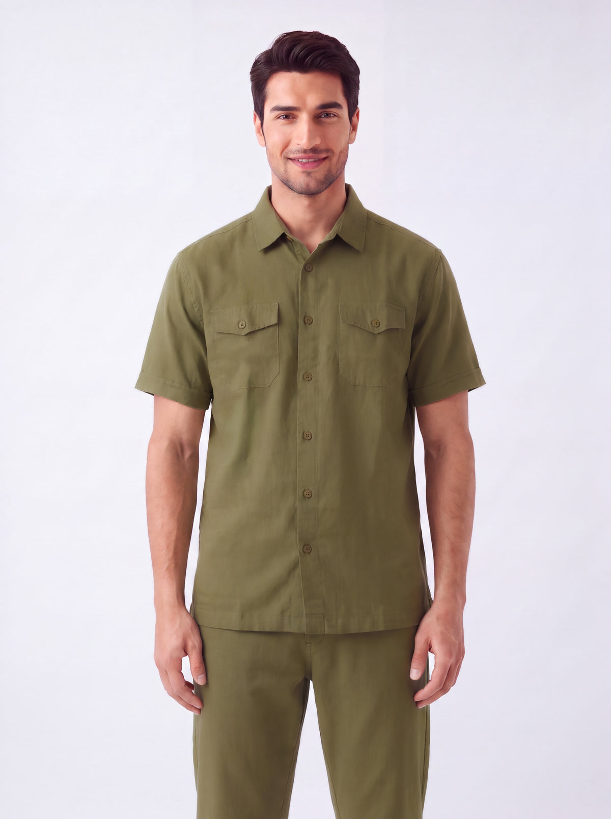 A man wearing a short-sleeve olive green button-up shirt and matching olive green pants, standing against a plain light gray studio background.
