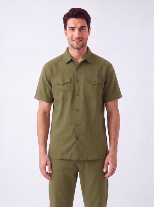 A man wearing a short-sleeve olive green button-up shirt and matching olive green pants, standing against a plain light gray studio background.