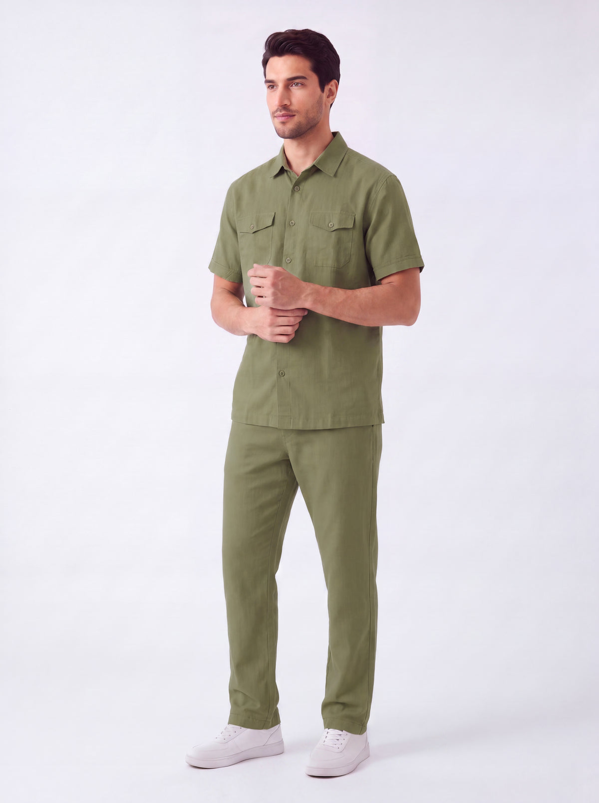 A man modeling a matching olive green short-sleeve button-up shirt and trousers set, paired with white sneakers, against a plain white studio background.
