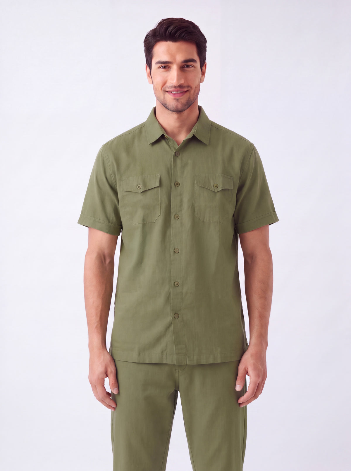 A man wearing a short-sleeve olive green military-style shirt and matching pants, standing against a plain light gray studio background.