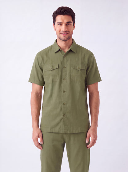 A man wearing a short-sleeve olive green military-style shirt and matching pants, standing against a plain light gray studio background.