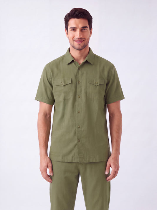A man wearing a short-sleeve olive green military-style shirt and matching pants, standing against a plain light gray studio background.