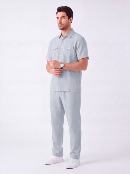 A man modeling a light gray short-sleeve button-up shirt and matching light gray trousers, paired with white sneakers, against a plain light gray studio background.