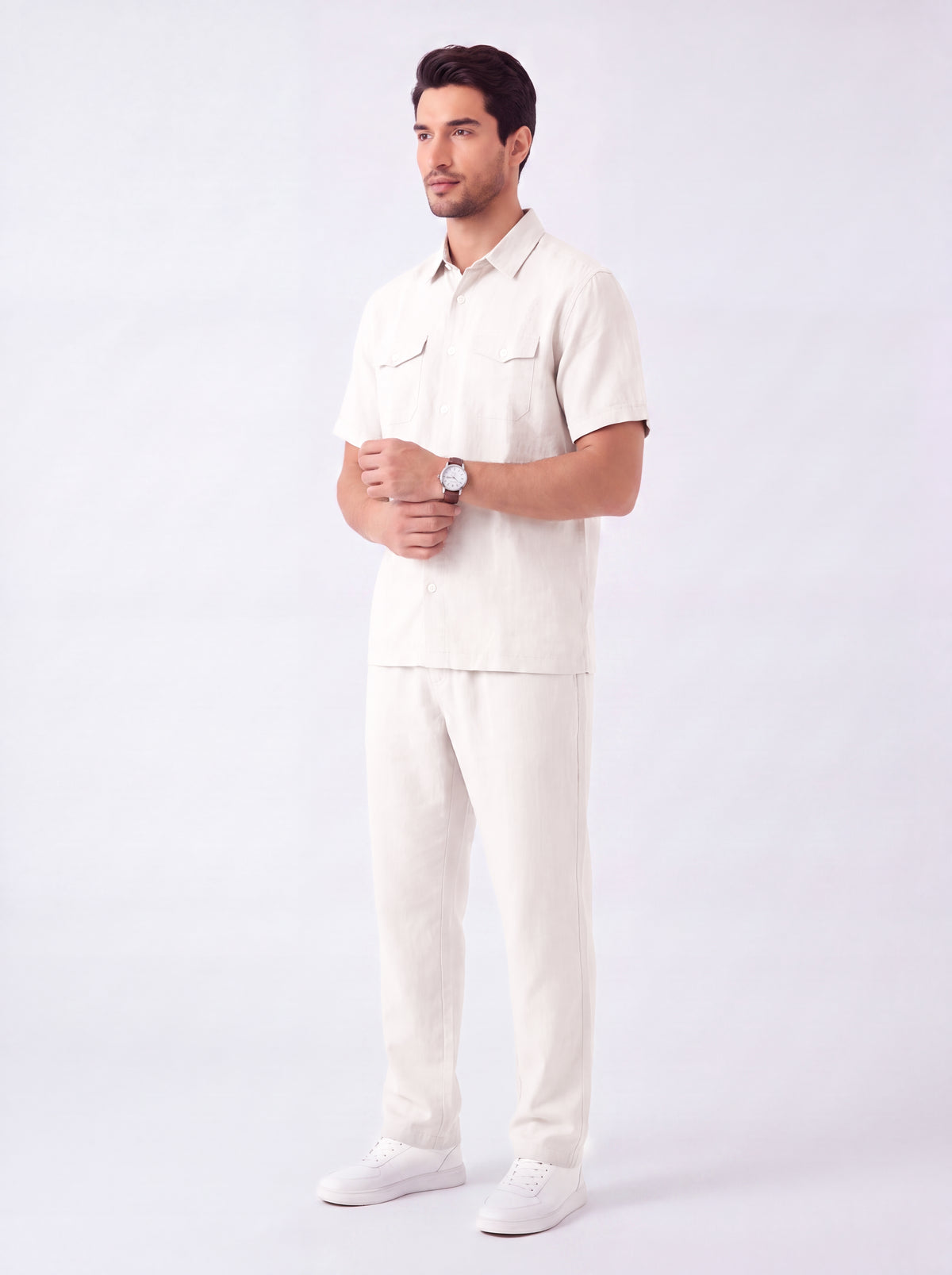 A man modeling a matching off-white short-sleeve button-up shirt and trousers set, paired with white sneakers, against a plain light gray studio background.