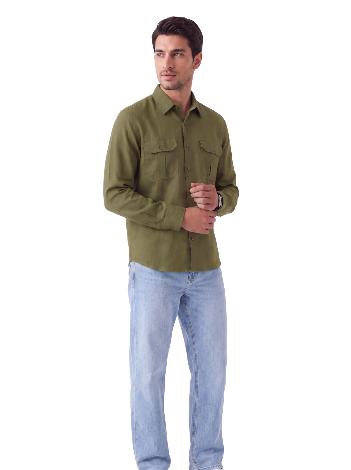A man wearing an olive green long-sleeve button-down shirt with two chest pockets and light blue jeans, standing against a plain white background.