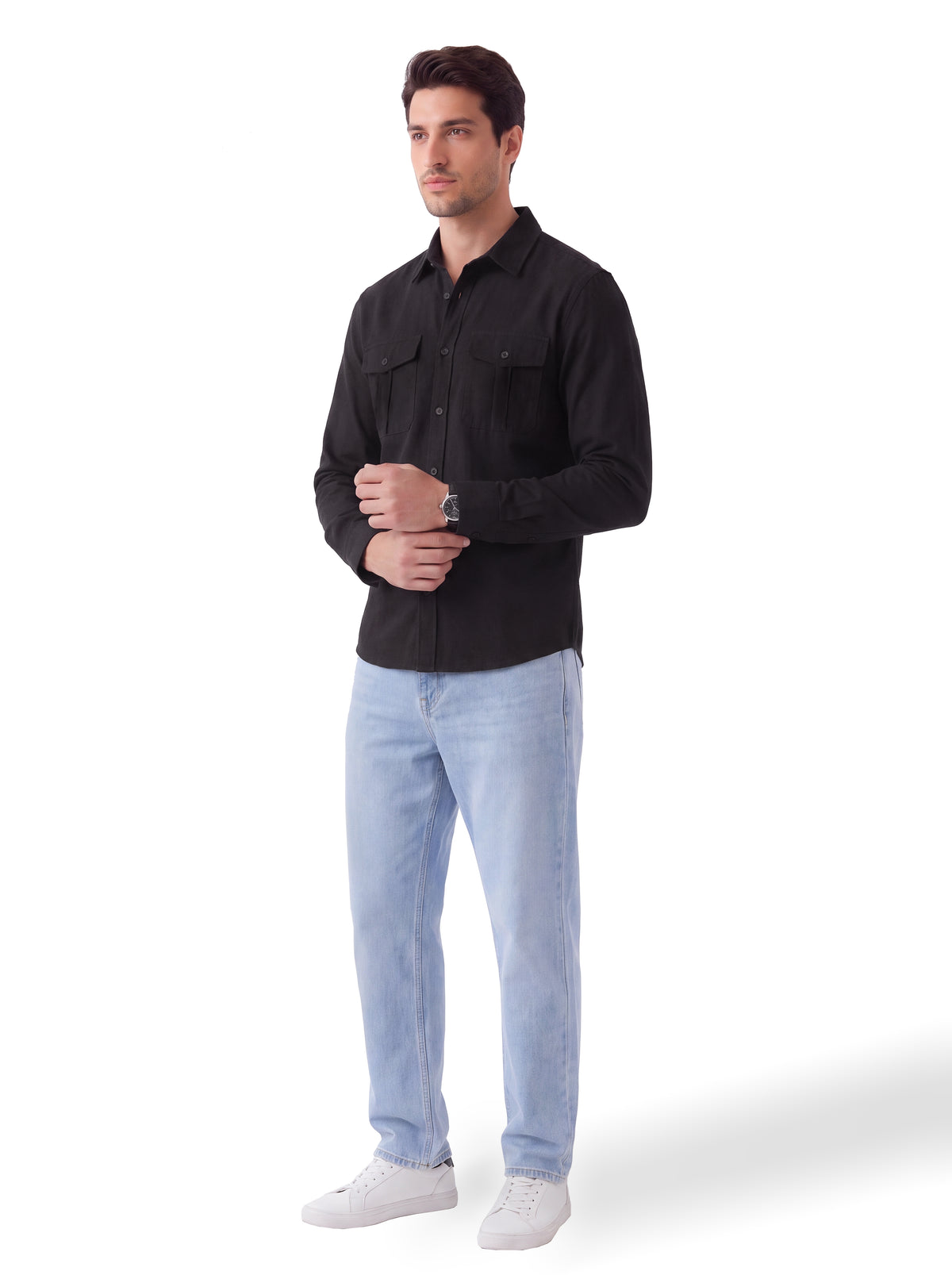 A man modeling a black long-sleeve button-up shirt with two chest pockets, paired with light blue jeans and white sneakers, against a plain white background.