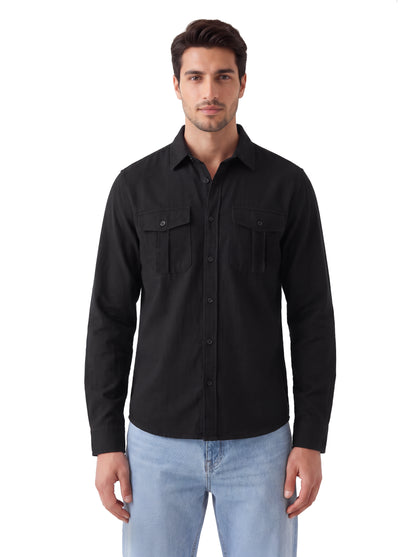 A man wearing a black long-sleeve button-up shirt with two chest flap pockets and light blue jeans, standing against a plain white background.