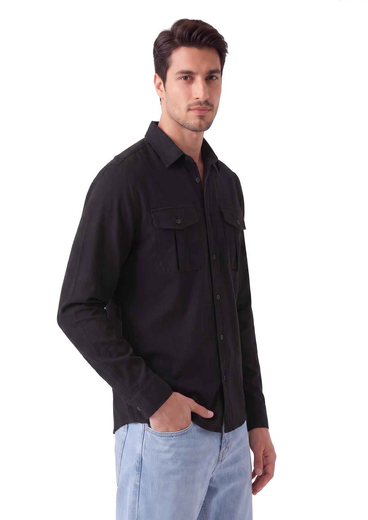 A man wearing a black long-sleeve button-up shirt with two chest pockets and light blue jeans, standing against a plain white background.
