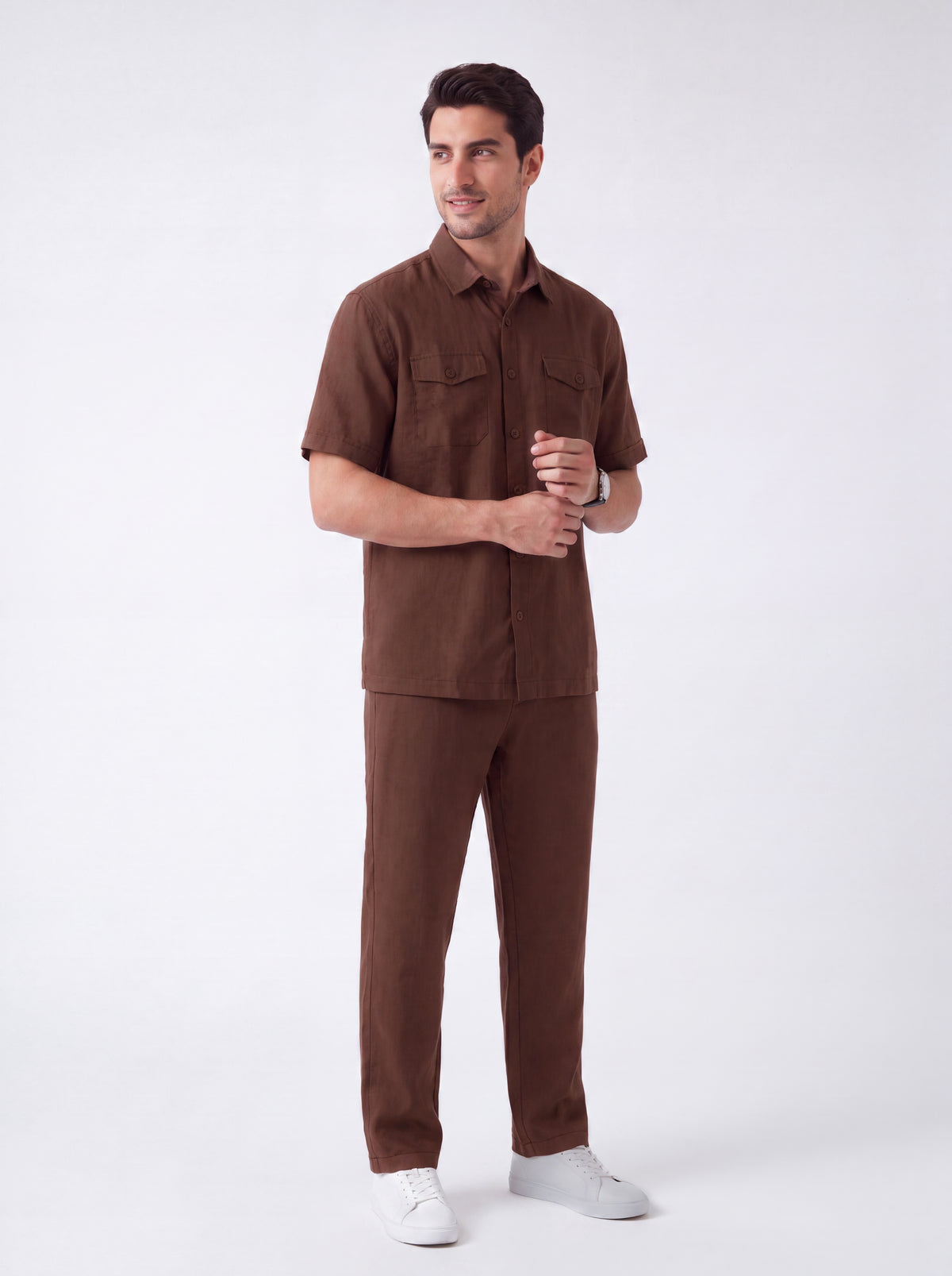 A man modeling a matching brown short-sleeve button-up shirt and trousers set, paired with white sneakers, against a plain light gray studio background.