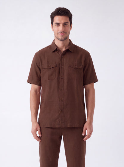 A man modeling a brown short-sleeve button-up shirt with two chest flap pockets, paired with matching brown trousers, against a plain light gray studio background.