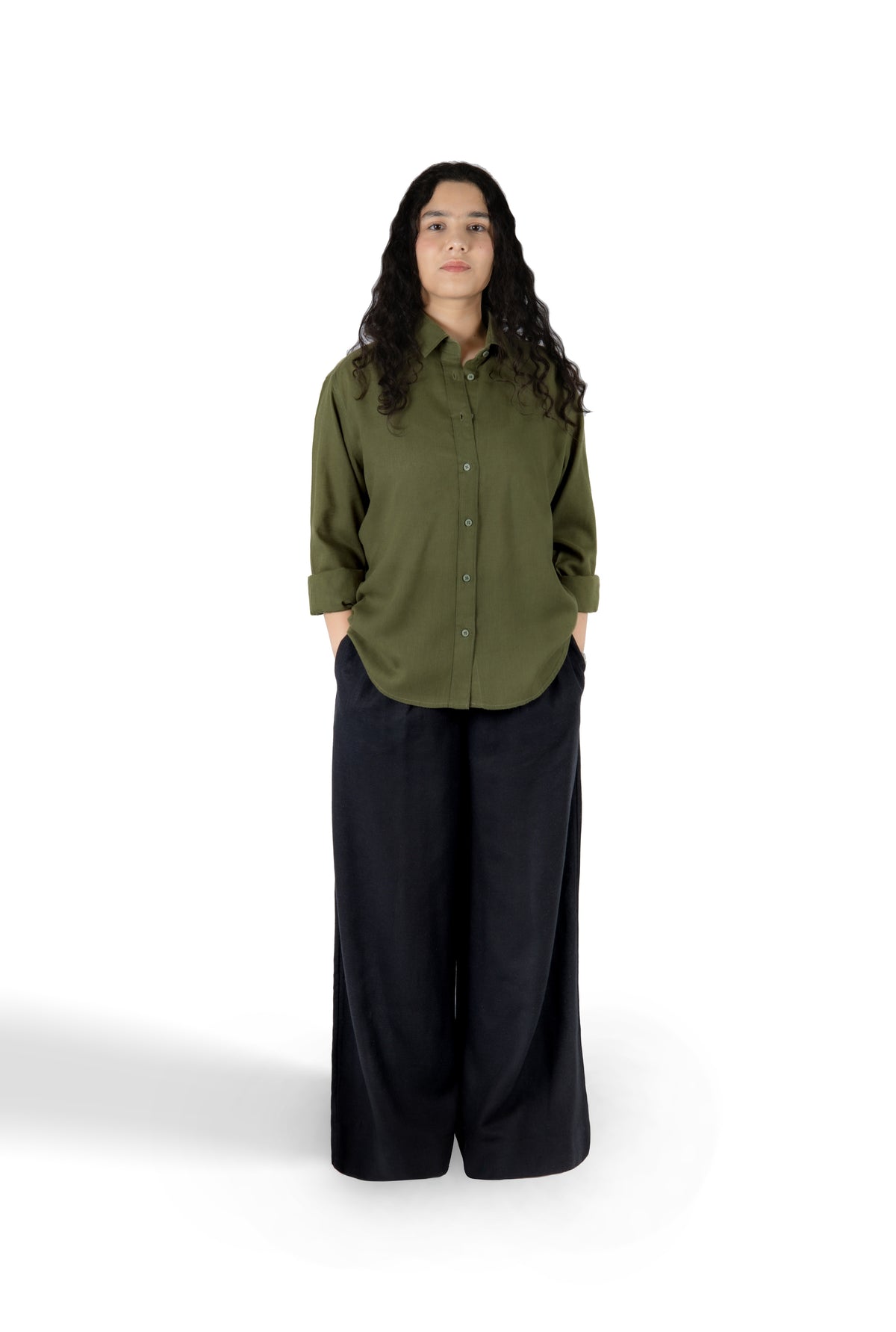 A woman stands against a plain white studio background wearing an olive green button-up shirt with dark wide-leg pants.