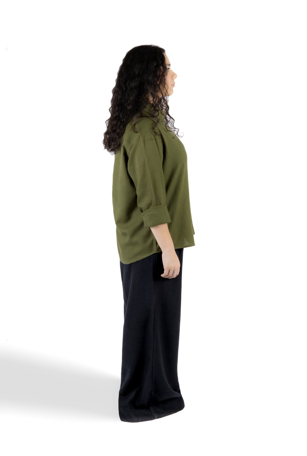 A woman shown in side profile wearing an olive green long-sleeve top and black wide-leg pants against a plain white studio background.