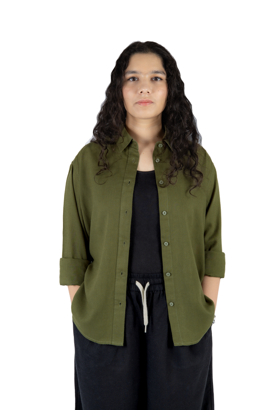 A woman with long, curly dark hair stands against a solid white background wearing an olive green button-up shirt over a black top and black pants.