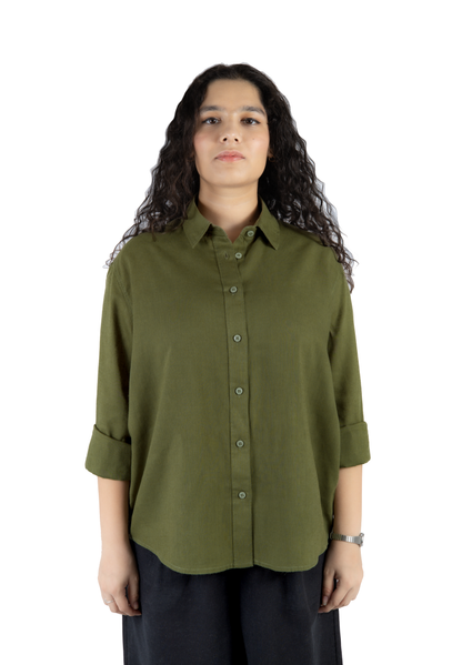 A woman with long, curly dark hair wearing an olive green button-down shirt with rolled-up sleeves and dark pants, standing against a plain white background.