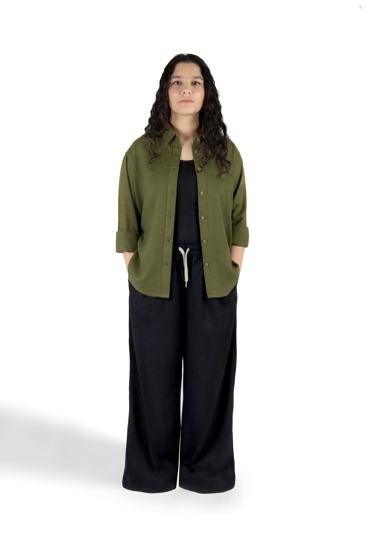 A woman stands against a white studio background wearing an olive green button-up shirt with three-quarter sleeves over a black top and black wide-leg pants.