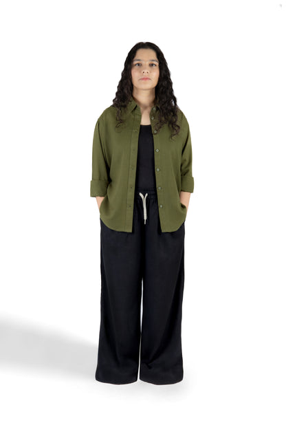 A woman stands against a white studio background wearing an olive green button-up shirt with three-quarter sleeves over a black top and black wide-leg pants.