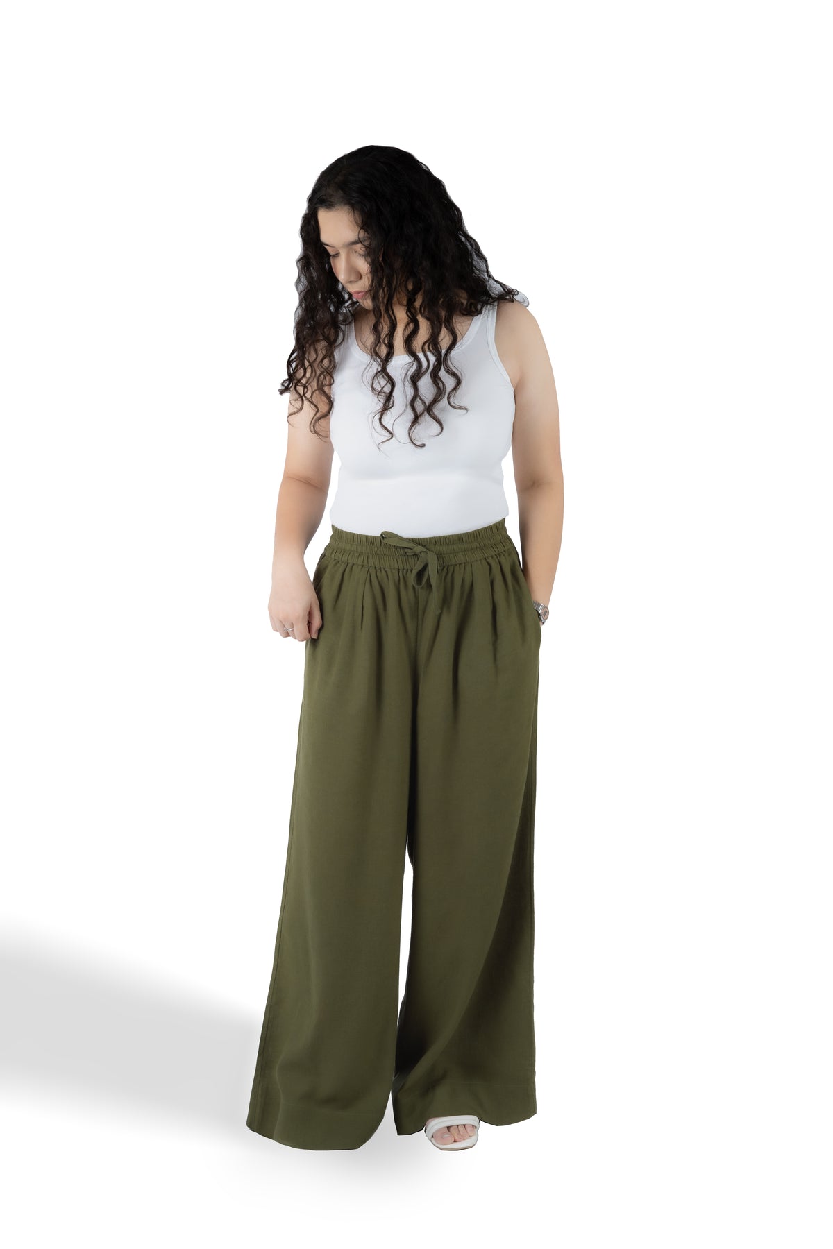 A woman modeling a pair of wide-leg olive green pants with a drawstring waist, paired with a white tank top, against a plain white background.