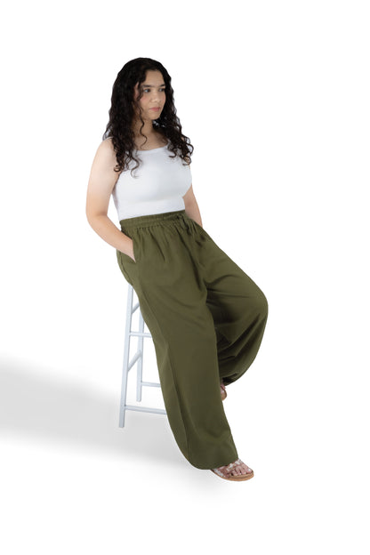 A woman with long dark curly hair sits on a white stool, wearing olive green wide-leg pants and a white sleeveless top against a plain white background.