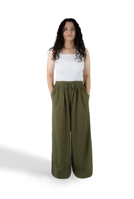 A woman with long curly dark hair stands against a white studio background wearing a white sleeveless top and olive green wide-leg pants.
