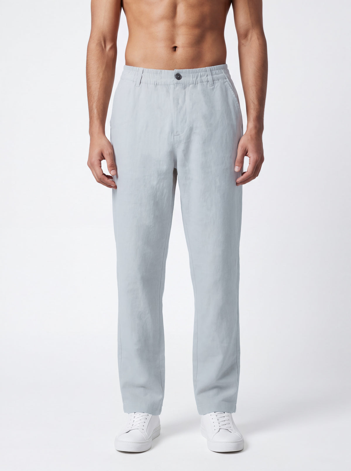 A man wearing light gray relaxed-fit trousers and white sneakers, standing against a plain light gray studio background.