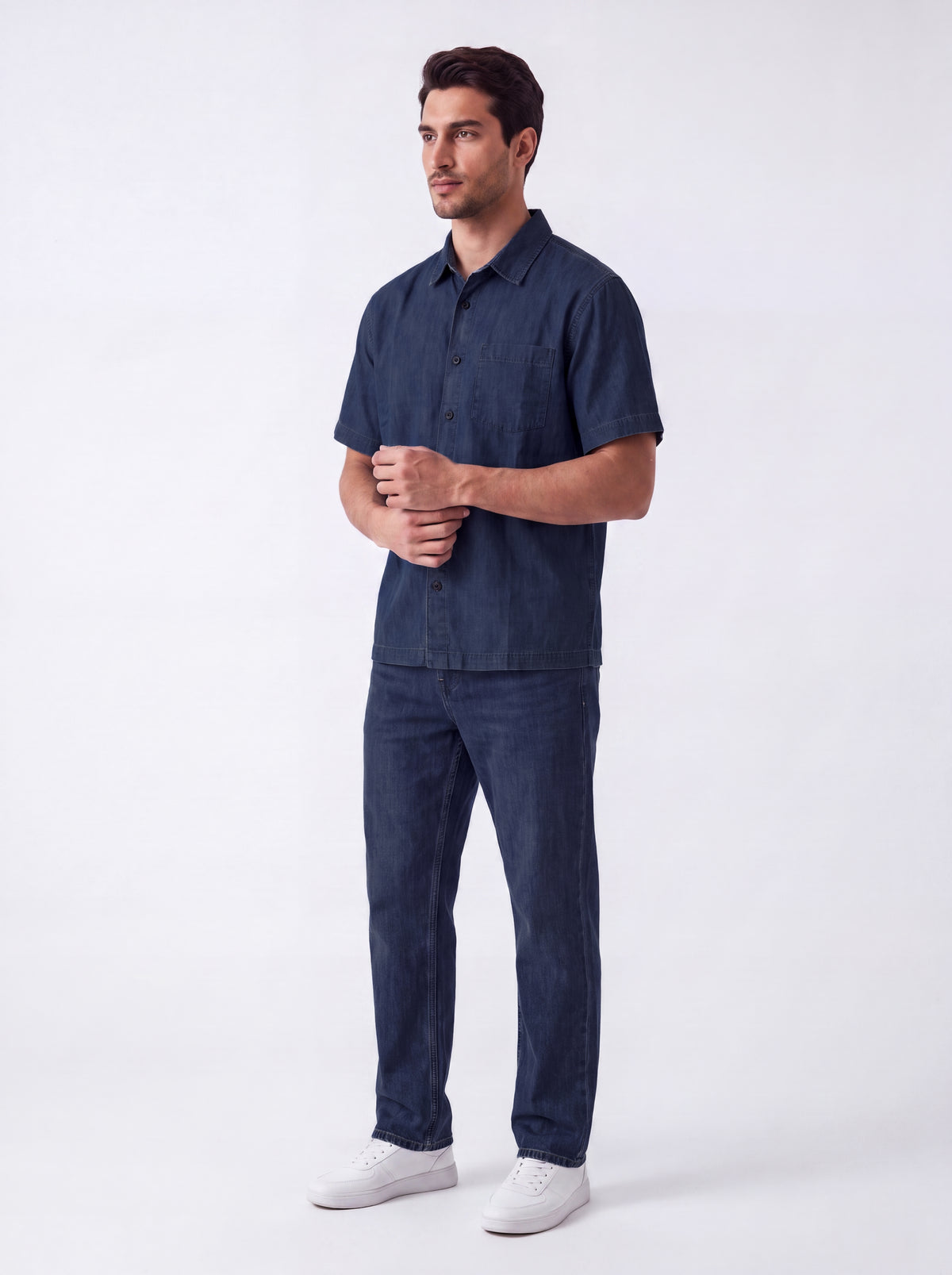 A man modeling a dark navy blue short-sleeve button-up shirt and matching navy blue jeans, paired with white sneakers, against a plain white studio background.
