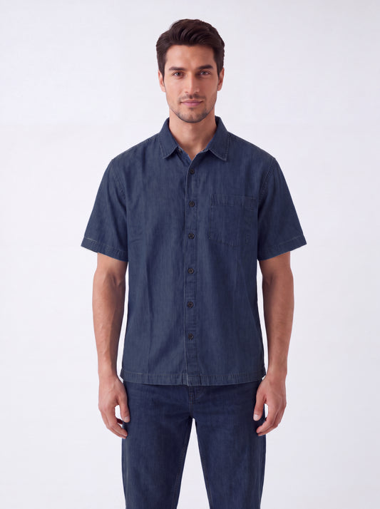 A man wearing a dark blue short-sleeve button-up shirt and matching dark blue jeans, standing against a plain light gray studio background.