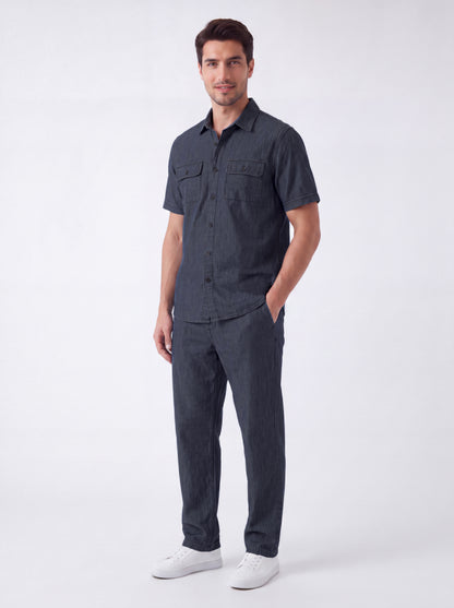 A man modeling a dark gray short-sleeve button-up shirt and matching dark gray trousers, paired with white sneakers, against a plain light gray studio background.
