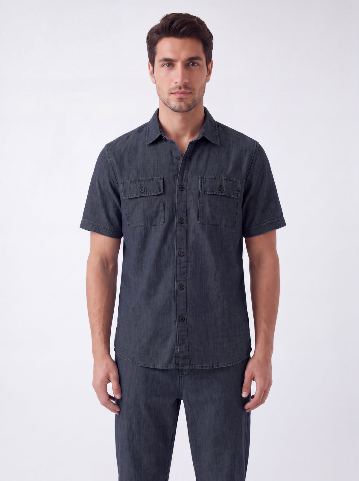 A man wearing a dark charcoal gray short-sleeve button-up shirt with two chest pockets, paired with matching dark pants, standing against a light gray studio background.