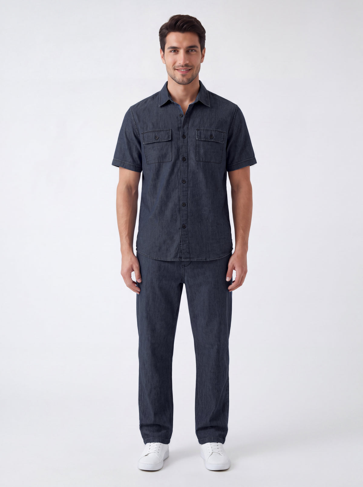 A man modeling a dark navy blue short-sleeve button-up shirt and matching dark navy pants, paired with white sneakers, against a plain light gray studio background.