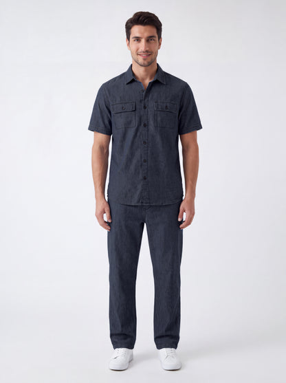 A man modeling a dark navy blue short-sleeve button-up shirt and matching dark navy pants, paired with white sneakers, against a plain light gray studio background.