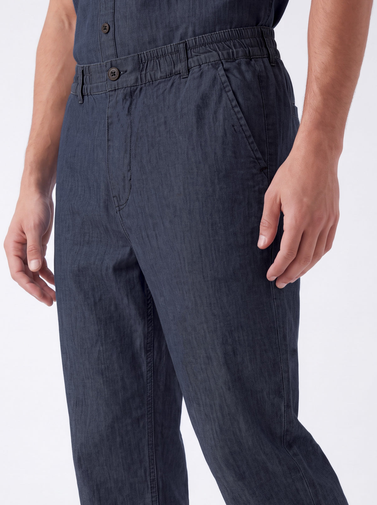 Close-up of a person wearing dark navy blue trousers, showing the waistband, front buttons, side pocket, and hands against a plain light background.