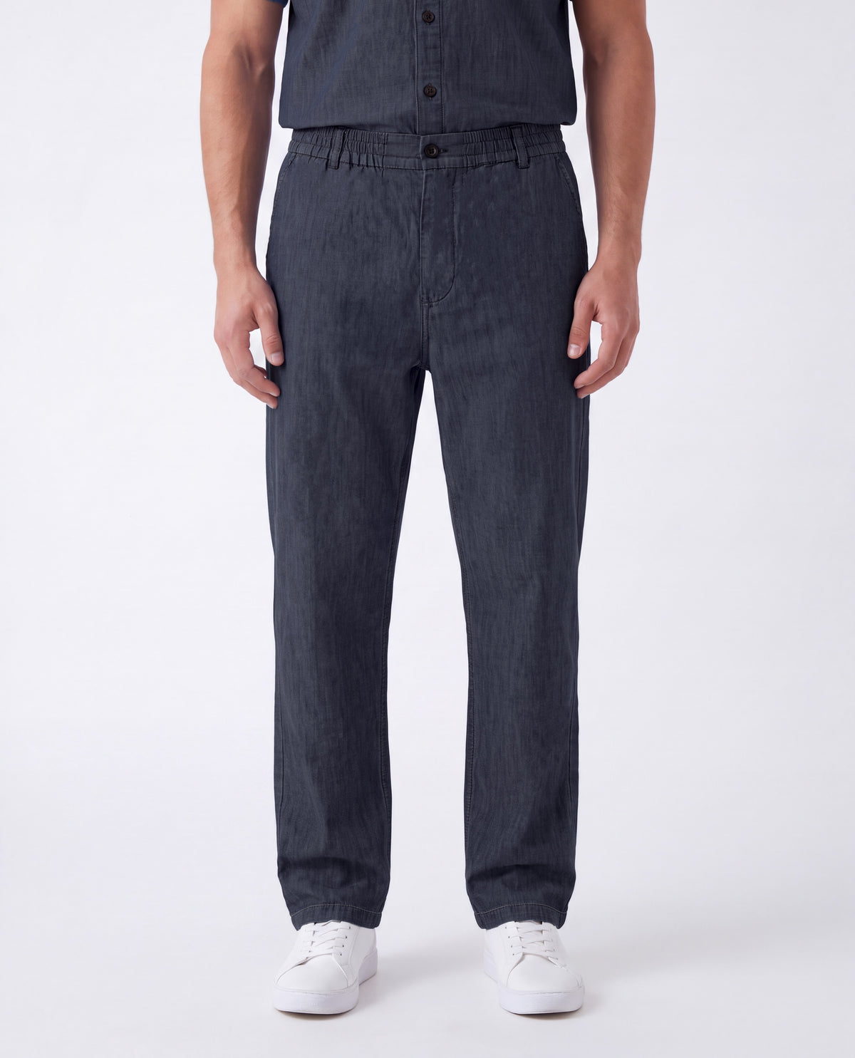 A person wearing dark charcoal gray slim-fit jeans with a button fly and side pockets, paired with a matching dark shirt and white sneakers, photographed against a clean white studio background.