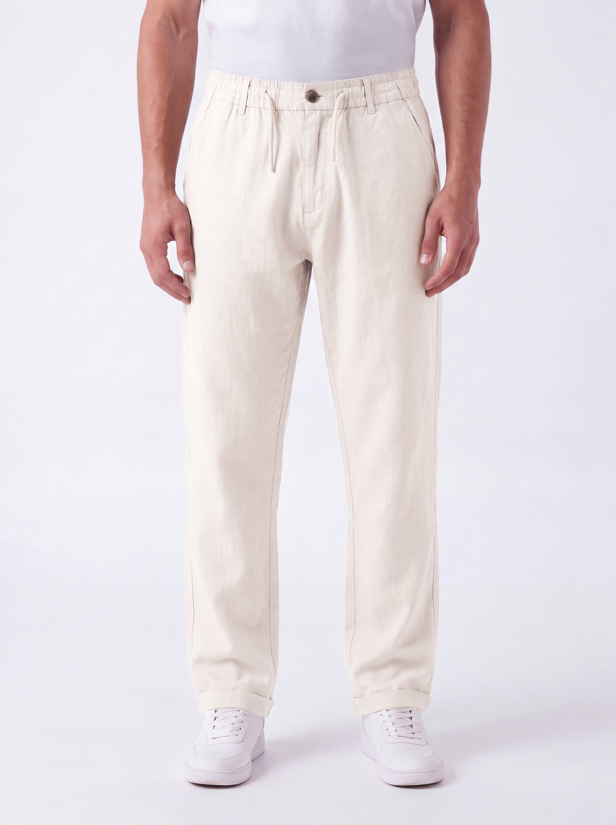 A person wearing off-white or cream-colored straight-leg trousers, paired with a white t-shirt and white sneakers, standing against a plain white studio background.