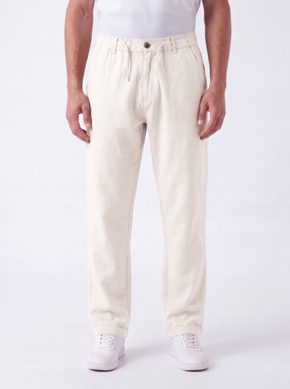 A person wearing off-white or cream-colored straight-leg trousers, paired with a white t-shirt and white sneakers, standing against a plain white studio background.
