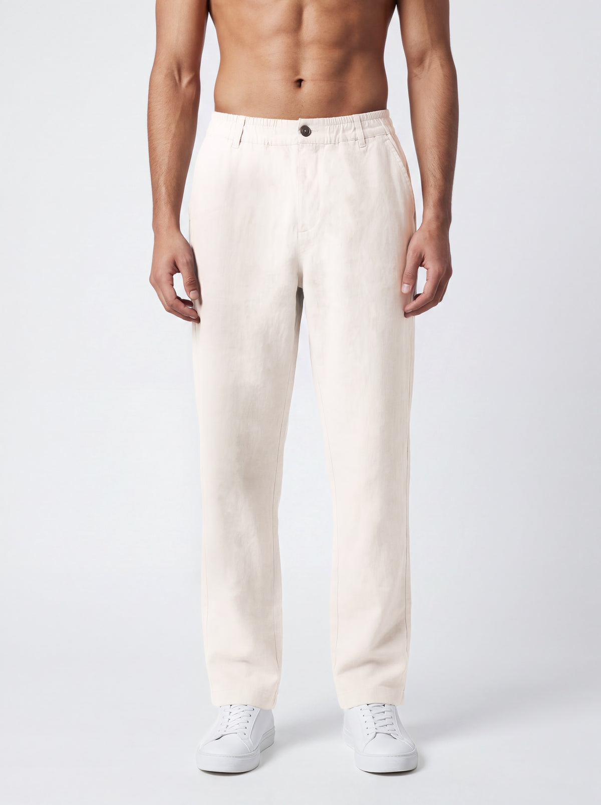 A man wearing off-white linen trousers and white sneakers, shown from the waist down against a light gray studio background.