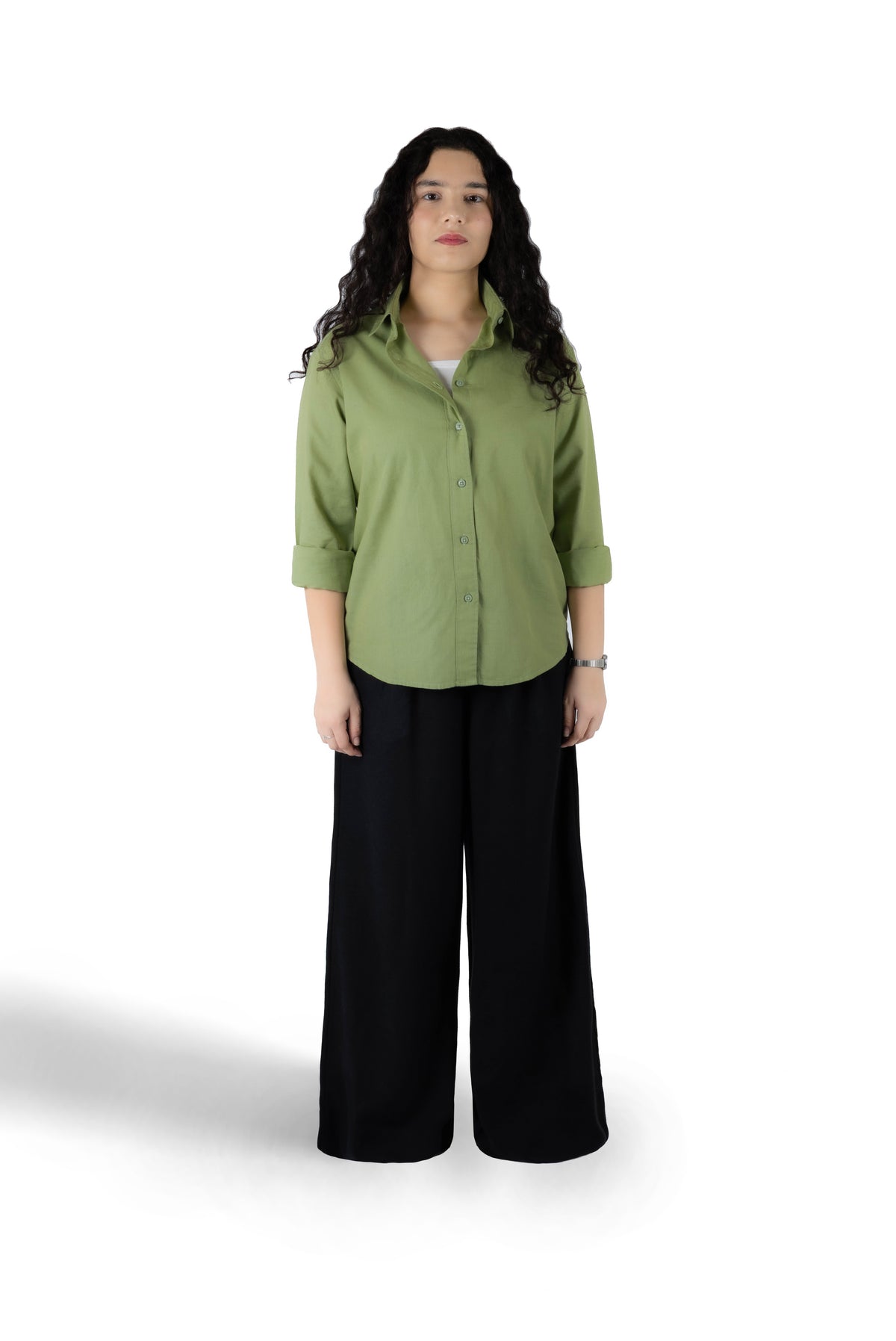 A woman stands against a white studio background wearing a muted sage green button-up shirt with rolled sleeves and black wide-leg pants.