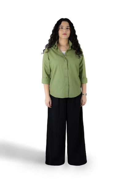 A woman stands against a white studio background wearing a muted sage green button-up shirt with rolled sleeves and black wide-leg pants.