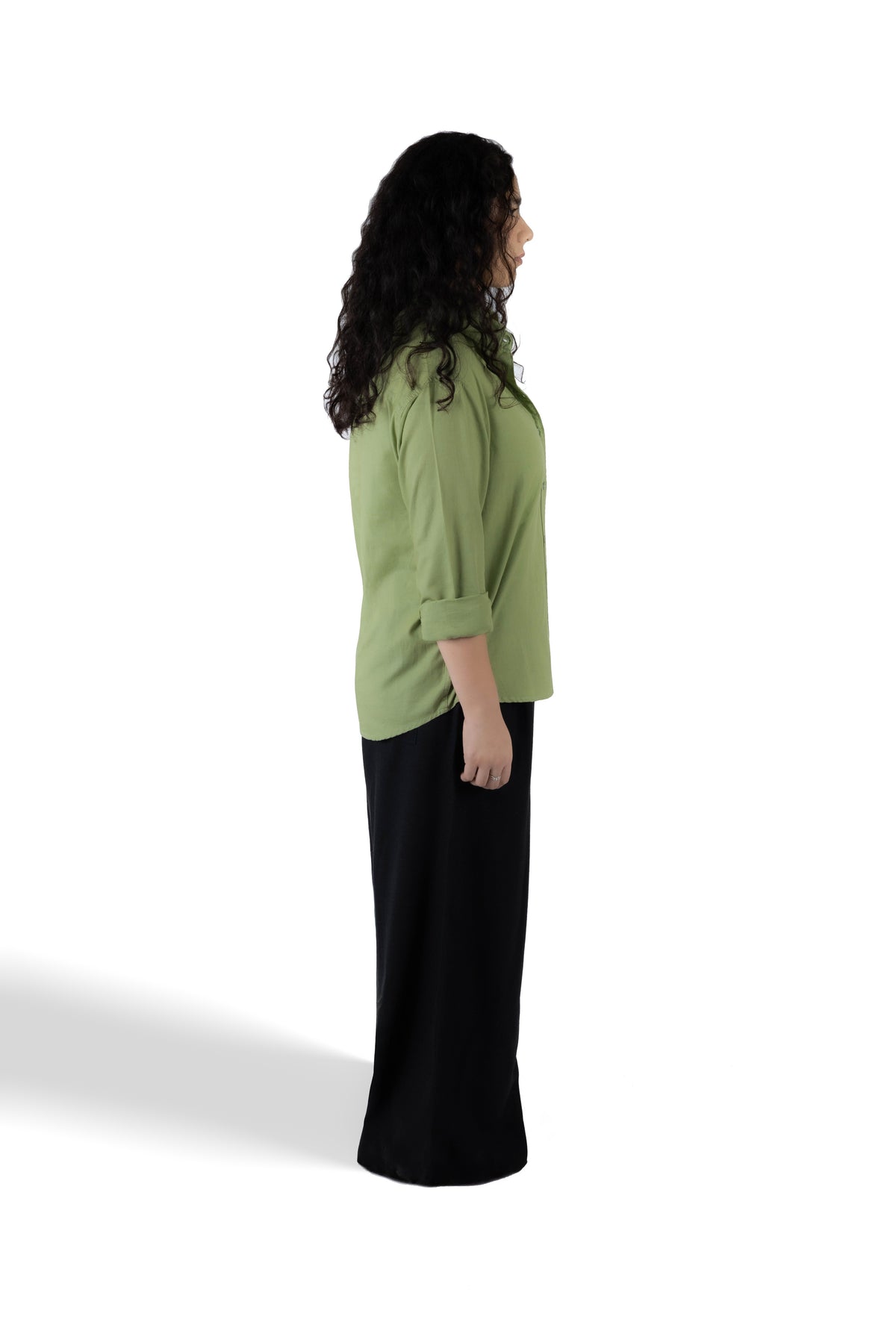 A woman shown from the side profile wearing a sage green three-quarter sleeve top and a long black skirt, standing against a plain white studio background.