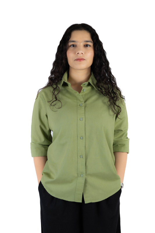 A woman with long, curly dark hair wearing a sage green button-down shirt with rolled-up sleeves and black pants, standing against a solid white background.